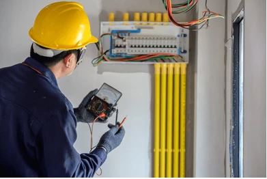 The image shows an electrician wearing a yellow hard hat using a digital testing device to inspect or troubleshoot an electrical panel with yellow conduit pipes and various circuit breakers.