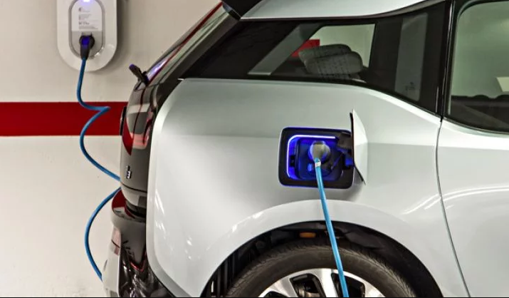 The image shows a white electric vehicle being charged in a garage, with a blue charging cable connected from a wall-mounted charging station to the car's illuminated charging port.