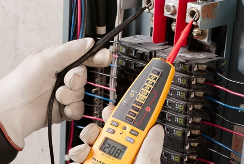 The image shows a technician's hands using a yellow digital multimeter with test probes to measure electrical readings on a circuit breaker panel, displaying "208" on the meter's screen.